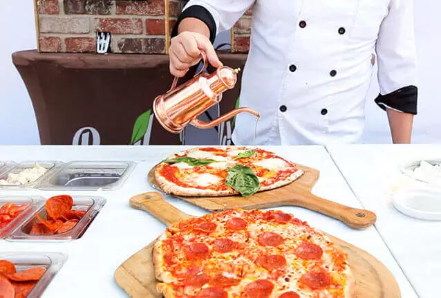 Chef drizzling olive oil over a fresh Margherita pizza beside a pepperoni pizza on wooden peels — Basil Pizza Bar, sister kitchen in the Bread and Butter Catering Group